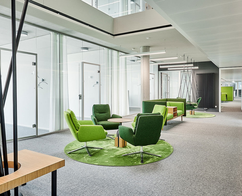 Moderne Büro-Lounge im Firmensitz von Medice, ausgestattet mit grünen Sesseln und Sofas von Brunner, Teppichen in Grasoptik und umgeben von Glastrennwänden.